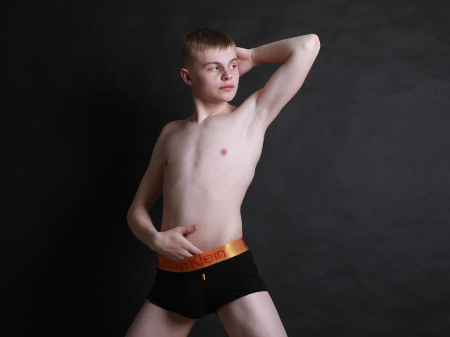HarleyQuinXO, shirtless, slim with pale skin, small nipples, short brown hair, black underwear with orange waistband, hand on hip, other arm behind head, dark background, confident pose.