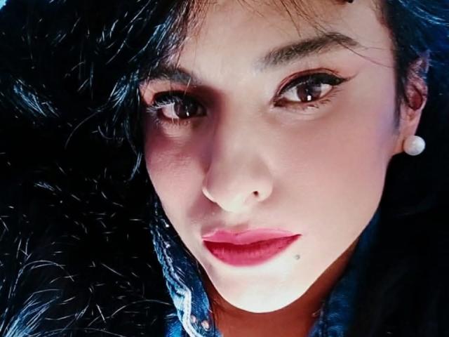 MoserratRouse has a cute face with dark wavy hair, red lipstick, and a small beauty mark below her right eye. She's wearing white earrings and has bold eyeliner.