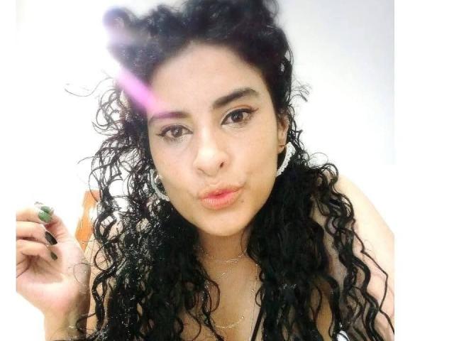 MoserratRouse with curly black hair, pouty lips, and green nail polish, holding a brush in her right hand, looking at the camera with a slight smile.