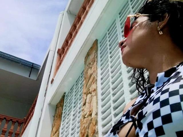 MoserratRouse with curly black hair, wearing checkered shirt, red sunglasses, and gold earrings, standing next to white shuttered windows and stone wall. Her tits are visible.