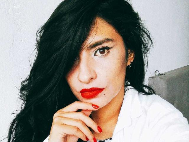 MoserratRouse with black hair, red lipstick, and red nail polish, wearing a white shirt, has her hand near her face, looking at the camera. Brown eyes, subtle makeup.