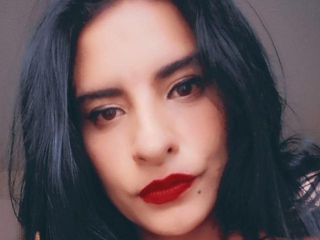 MoserratRouse's face, featuring dark, wavy hair, red lipstick, and a small beauty mark under her right lip, with brown eyes and a slight smirk.