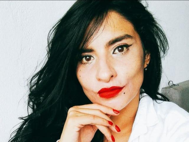 MoserratRouse has dark, wavy hair, red lipstick, and red nail polish, with a white shirt and brown eyes, looking at the camera with a slight smile.