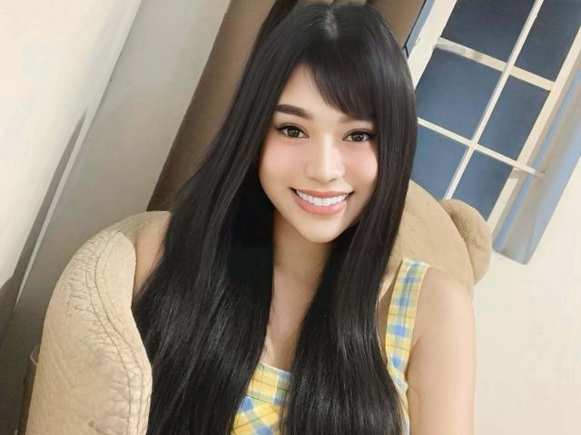 TransAsianSweetX with long black hair, smiling, wearing a yellow plaid dress, sitting in a beige chair, has cute brown eyes and a small nose, and is attractive.