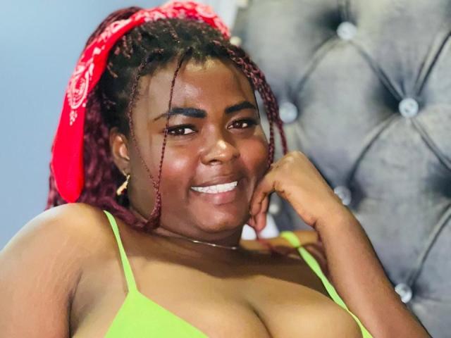 KarlaFlorez with dark skin, red braided hair in a red bandana, smiling, green bra, hand on cheek, gray tufted headboard, earrings, showing her tits, looking cute.