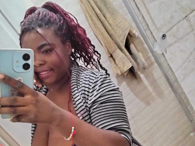 KarlaFlorez with red curly ponytail, smiling, taking selfie in bathroom mirror. She's wearing a striped shirt, showing her tits, holding a blue phone, and has a red bracelet.