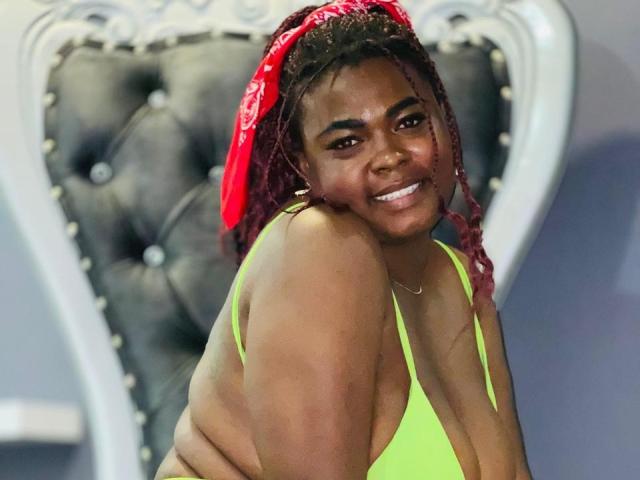 KarlaFlorez with dark skin, red headscarf, and pink braids, wearing a neon green bikini, smiling with her ass and tits slightly visible, sitting on a black tufted chair.