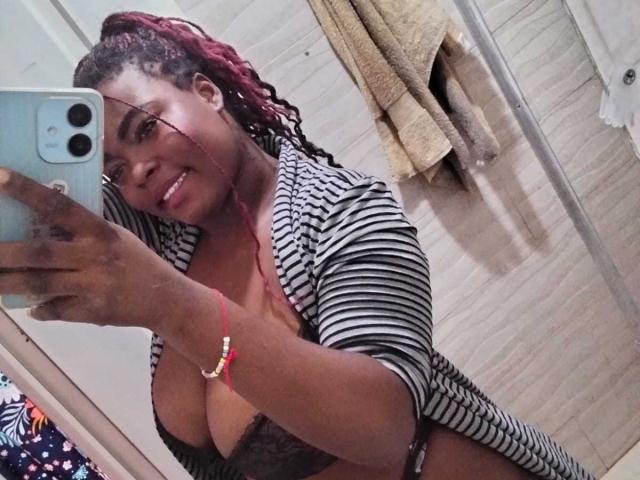 KarlaFlorez with dark skin, red and black braided hair, striped shirt, black lace bra, smiling, holding phone, mirror selfie, visible cleavage, red bracelet, towel in background.