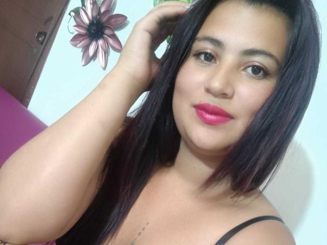 LesbianFourCouple with dark brown hair, pink lipstick, and black spaghetti straps, touching her head, smiling, tattoo below collarbone, light skin, brown eyes.