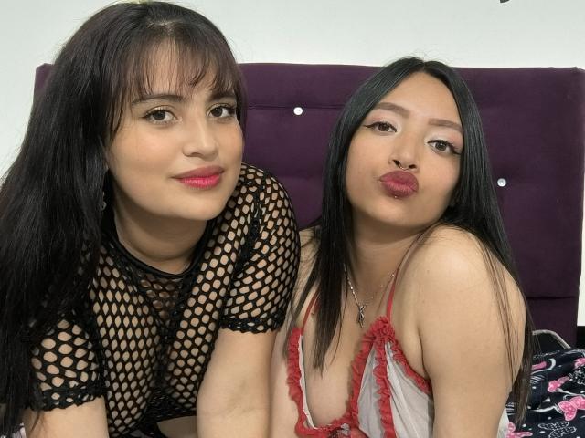 Two Asian women with long black hair, wearing fishnet and red lace lingerie, kissing boobs, looking at camera, pink lipstick, purple headboard in background, soft light, cute.