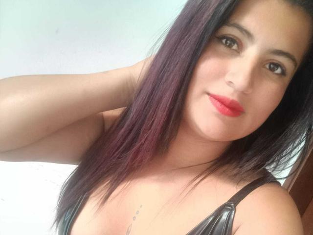 LesbianFourCouple with long dark purple hair, red lipstick, and black leather top, showing off small tits and a tattoo below her chest, smiling at the camera.
