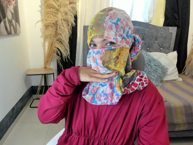 AniisaAli wearing a colorful, patterned hijab and red shirt, covering her mouth with her hand. Her eyes are visible, and she has a pensive expression.