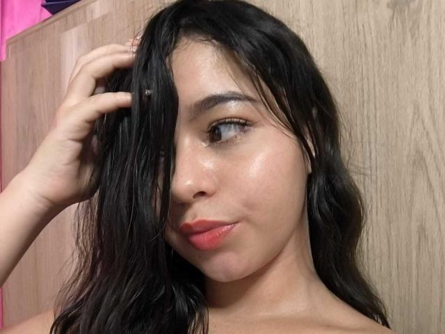 LissyCarter has shiny brown skin, black wavy hair, and red lipstick. She's touching her hair with one hand, showing a subtle smile. Her tits are not visible.