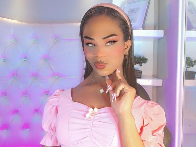 DarlingSweety with long black hair, pink headband, and puffy pink dress, touching her chin with manicured nails, wearing a heart necklace, and pink eyeshadow.