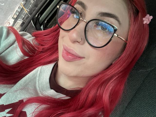 SammyViolet with red hair, glasses, nose ring, and pink lips, wearing a grey sweater with a red V-neck, sitting in a car. Her pussy is not visible.