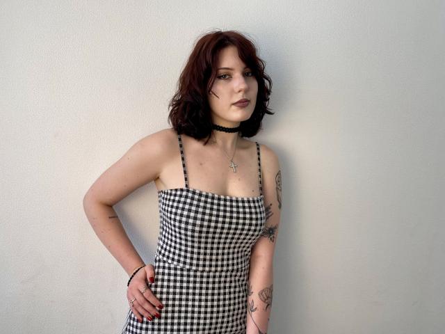 Khori with wavy red hair, black check dress, strap tattoos, choker, hands on hips, subtle makeup, looking at camera, pale skin, small tits, cross necklace.