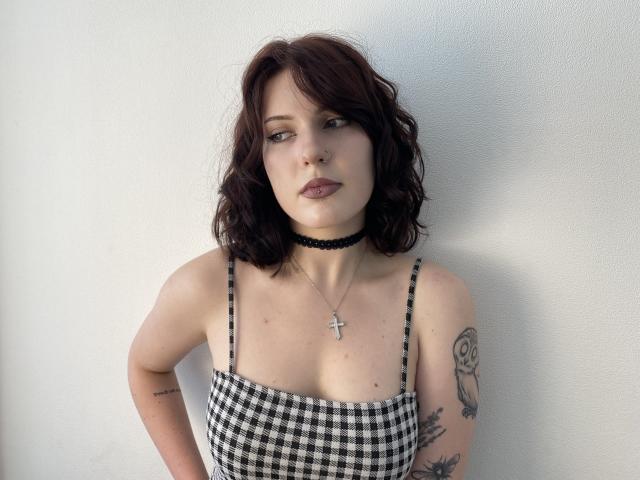 Khori with wavy dark brown hair, wearing a black and white checkered dress, black choker, and cross necklace, showing tattoos on her arms, and looking slightly to the side.