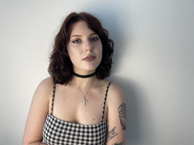 Khori with wavy brown hair, wearing a black choker and checkered top, has tattoos on her arm and wears a cross necklace, staring with a neutral expression.