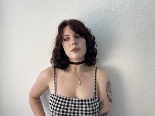 Khori has wavy dark brown hair, a black choker, checkered top with straps, small tits, tattoos on arms, and a cross necklace, standing against a white wall.