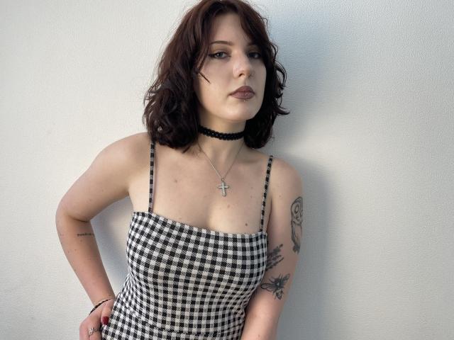 Khori with shoulder-length brown wavy hair, black checkered dress, black choker, cross necklace, tattoos on left arm, standing against white wall, looking slightly to the side.