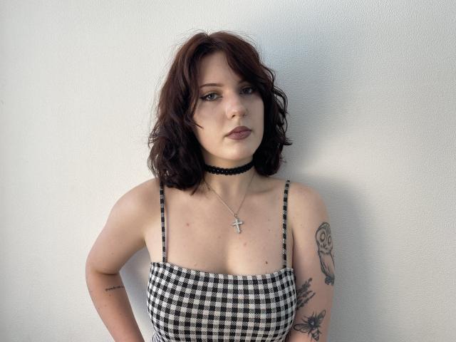 Khori with wavy brown hair, black choker, checkered top, small cross necklace, tattoos on arm, and serious expression, showing her tits and a slightly visible ass.