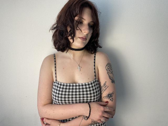 Khori with wavy brown hair, black choker, checkered tank top, showing off her tits and tattoos, arms crossed, cross necklace, pale skin, red nails, white wall background.