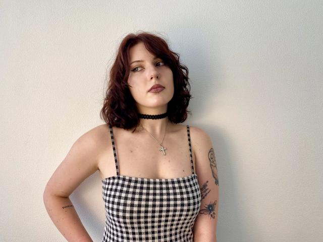 Khori with wavy red hair, black choker, checkered tank top, showing small tits, tattoos on arms, hands on hips, looking slightly up, neutral expression, white wall background.