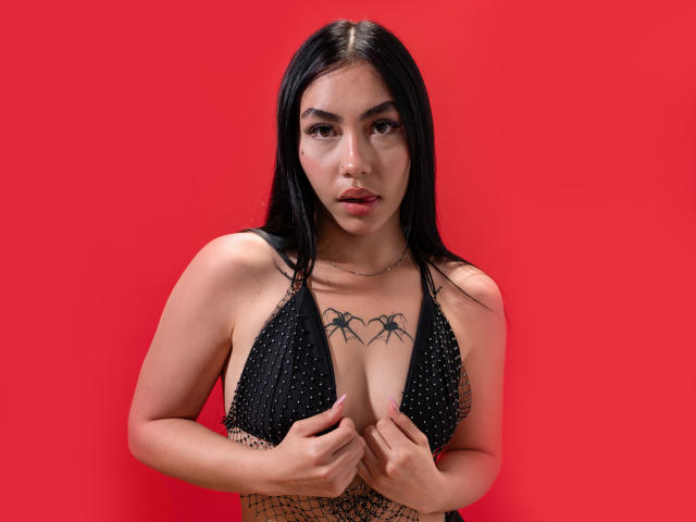CelesteConner, con cabello negro, vistiendo un top de bikini negro con tachuelas, sosteniendo sus pechos, tatuaje de araña sobre sus pechos, fondo rojo, mirando directamente a la cámara con los labios entreabiertos..