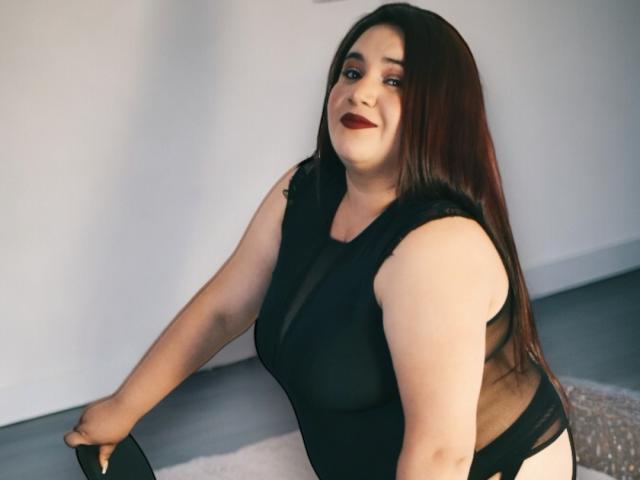 KhloeThompson has long brown hair, fair skin, and red lipstick. She's wearing a black sheer top, revealing her large boobs, and black panties. She's sitting with a slight smile.