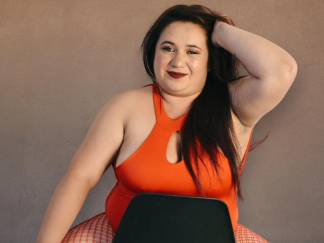 KhloeThomson with pale skin, dark brown hair, red lipstick, wearing an orange sports bra and red checkered pants, smiling, hand in hair, laptop on her lap.