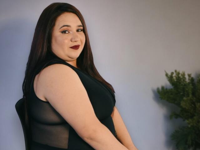 KhloeThomson with dark brown hair, wearing a black sleeveless dress, showing her tits through sheer fabric, looking over her shoulder with a slight smile, dark red lipstick.