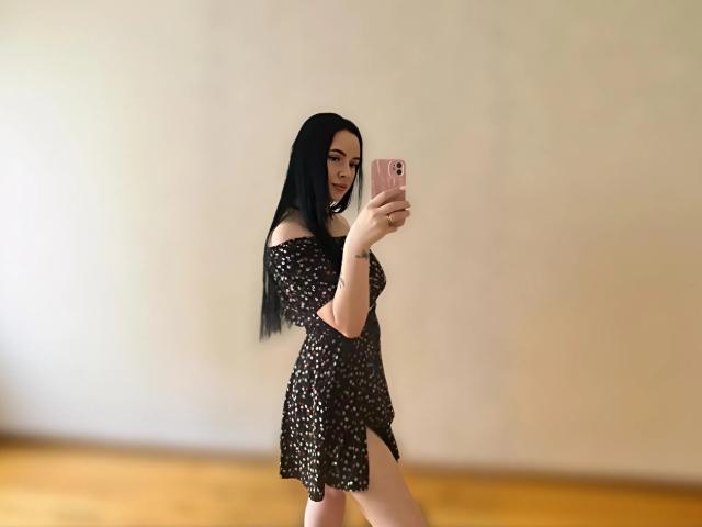 Lulii69 with long black hair, off-shoulder black dress with white dots, taking a selfie in a mirror, holding a pink phone, standing on wooden floor.