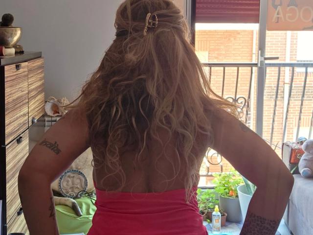 ZoeRubia-hot with wavy blonde hair in a ponytail, back facing camera, wearing a pink dress, showing tattoos on arms, standing by a balcony with potted plants.