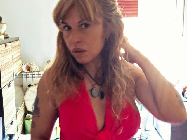 ZoeRubia-hot with blonde hair, wearing a red halter top, showing her big tits, looking at the camera, hand in hair, necklace, tattoo on left arm, in a sunlit room.