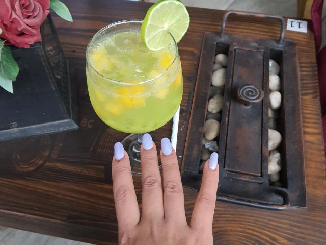 LizBonnet's hand with light blue nail polish holds a glass of green-yellow drink with ice and a lime slice, next to a rustic stone holder on a wooden table.