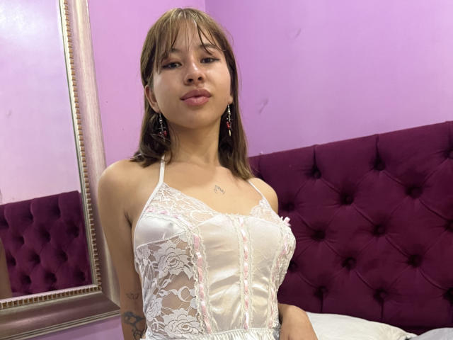 KattyyKate with small tits, wearing a white lace lingerie top, purple tufted headboard, brown hair, earrings, and a tattoo on her left arm, in a pink-walled room.
