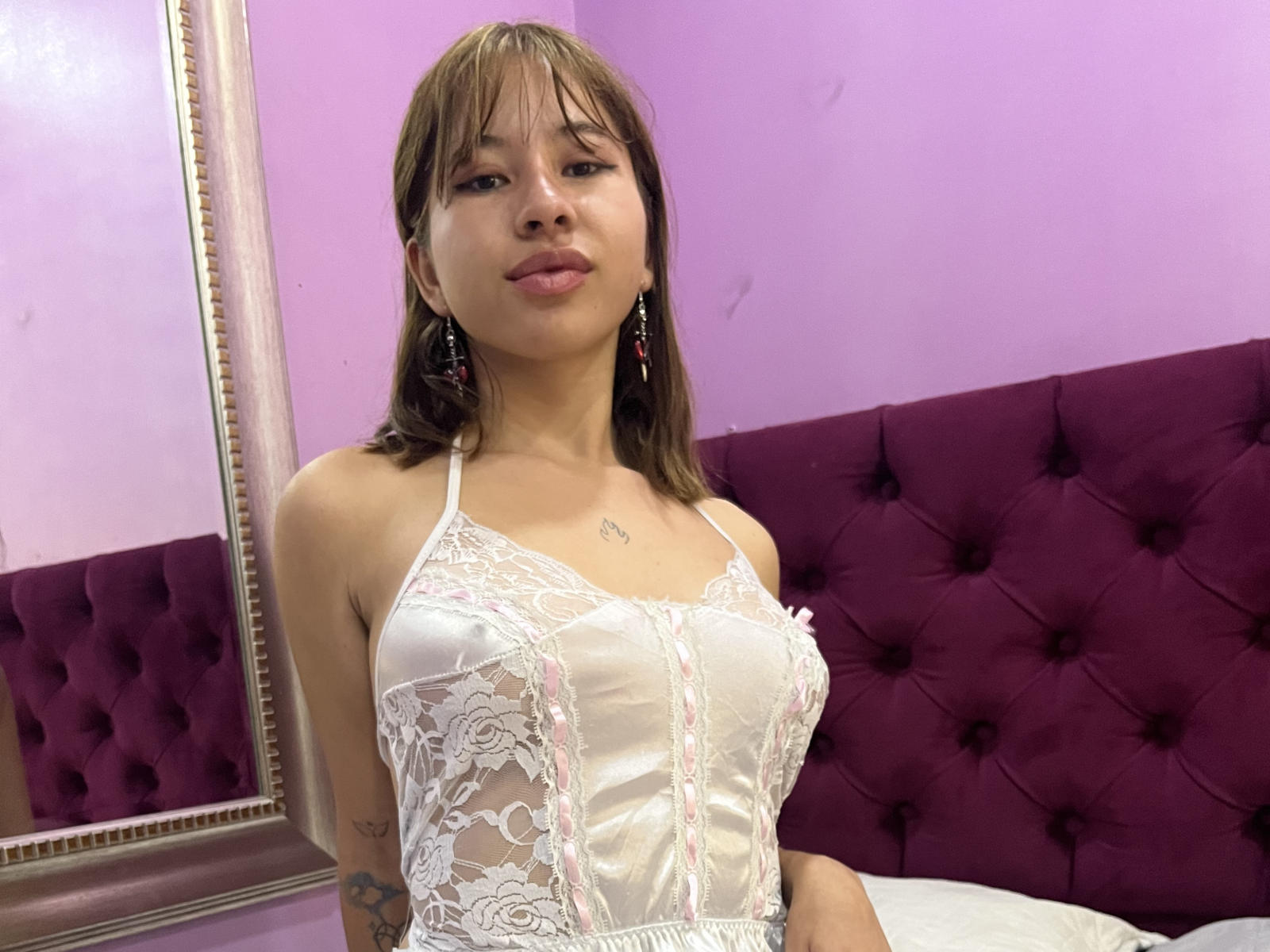 KattyyKate with small tits, wearing a white lace lingerie top, purple tufted headboard, brown hair, earrings, and a tattoo on her left arm, in a pink-walled room.