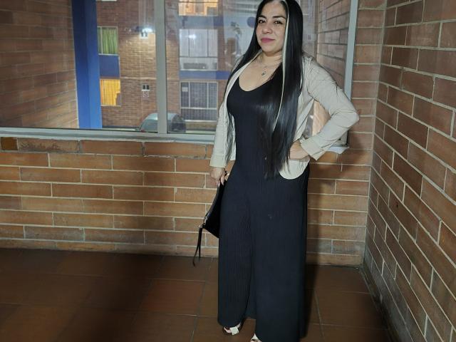 EvaLunaLatina with long black hair, wearing a black dress and beige cardigan, hands in pockets, standing against a brick wall, holding a black bag, smiling.