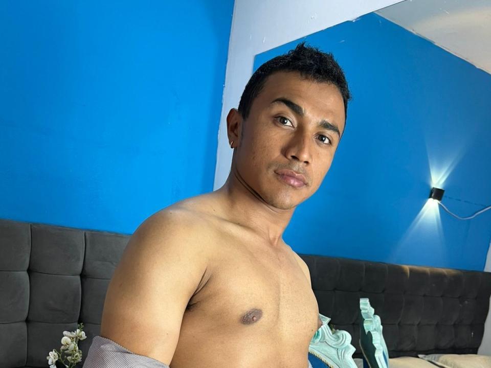 Shirtless OliverClarkk with dark brown skin, short black hair, and small nipples, standing in a blue room, looking at the camera with a slight smile.