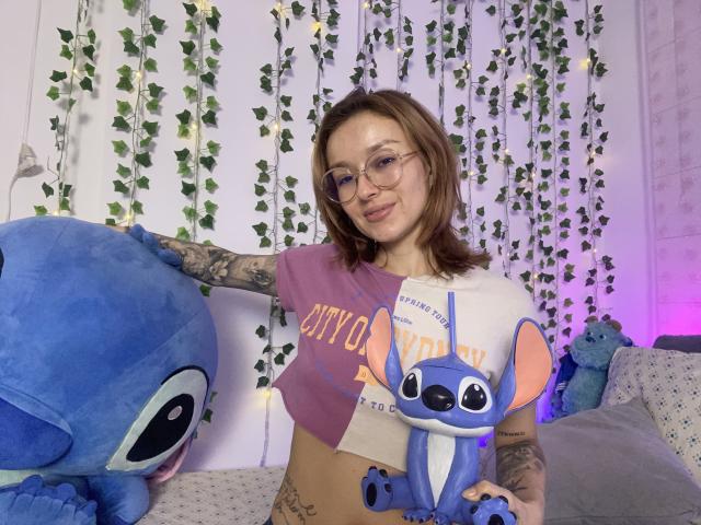 SarahRousen with glasses, tattoos, brown hair, and a split-shirt showing her tits, holds two Stitch plushies, one in her hand and one on her arm.