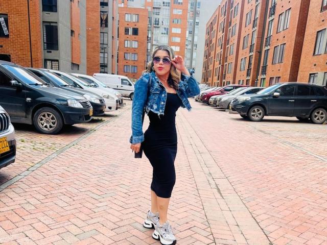 LiaRousselle with big ass, wearing a denim jacket, black dress, and sneakers, holding a phone, standing on a brick sidewalk, in front of parked cars and red brick buildings.