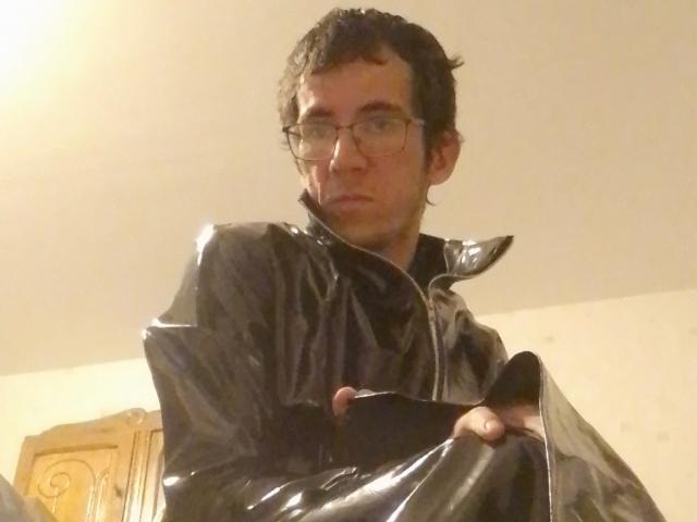 Bricebd, wearing a shiny black leather jacket, stands with a serious expression, holding a black object in front of him. He has glasses and curly dark hair.