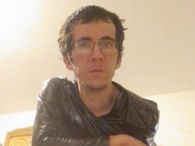 Bricebd has wet, dark hair and glasses, wearing a shiny black plastic jacket, looking slightly confused. His ass is not visible.