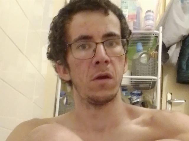 Shirtless Bricebd with curly brown hair, glasses, and a stubble, looking slightly surprised, in a bathroom with a metal shelf and toiletries in the background.