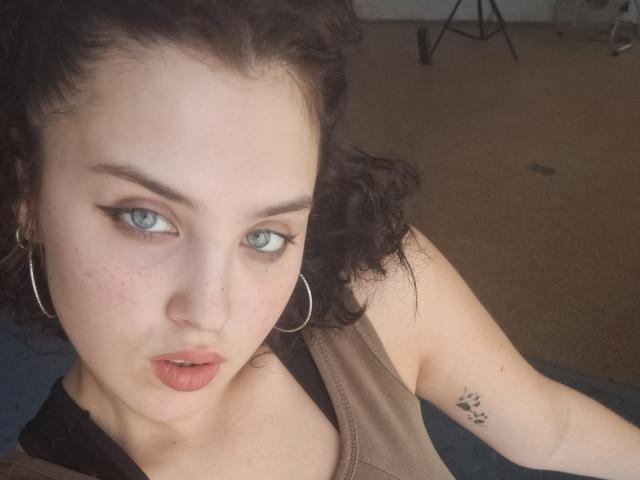 HeillyHot with curly black hair, pale skin, blue eyes, and hoop earrings, showing a small tattoo on her arm, pouty pink lips, and a brown sleeveless top.
