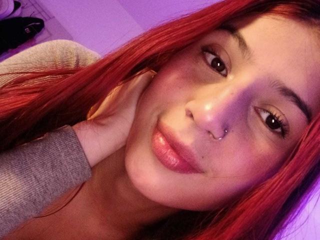 SophieBeaumont with red hair, nose ring, and lip ring, smiling slightly, hand on cheek, wearing gray long-sleeve shirt, in a purple-lit room.