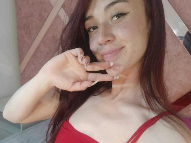 AmyKobold69 with long red hair, smiling, touching her pink-lipsticked bottom lip, wearing a red top, showing off manicured nails, with a cute, sexy, and attractive look.