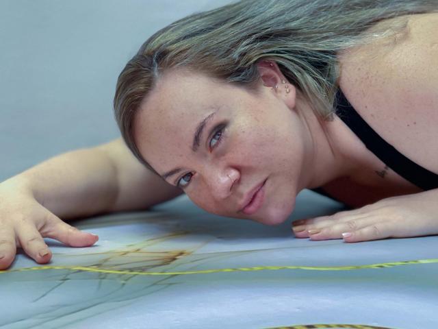 TalianaCediel is lying on her stomach with her face close to the camera, wearing a black bra. She has freckles, blue eyes, and blonde hair. Her hand is on a yellow line.