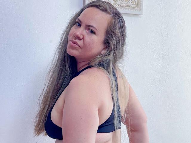 TalianaCediel with long, messy blonde hair, black bra, and pouty expression, showing her ass and tits, standing against a white wall, looking over her shoulder.