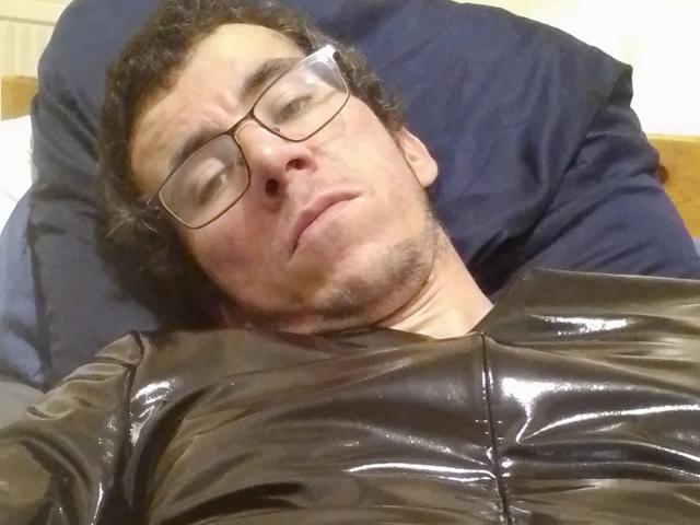Bricebd with glasses, curly dark hair, and a slight beard, wearing a shiny brown latex shirt, lying on a dark pillow, looking bored with a relaxed expression.